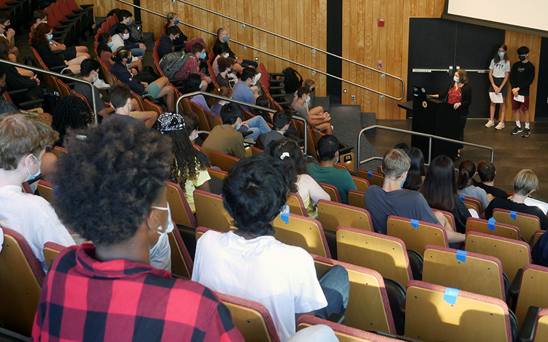 A photo from Upper School assembly with Upper School Head Neeltje Henneman at a podium, and students Ella Kulper and Daniel Hu standing behind her. The backs of students, seated in auditorium chairs, are in the foreground.