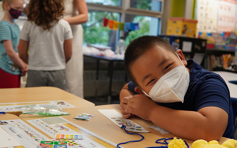 A Lower School student wearing a mask and smiling at the camera while seated at a classroom desk.