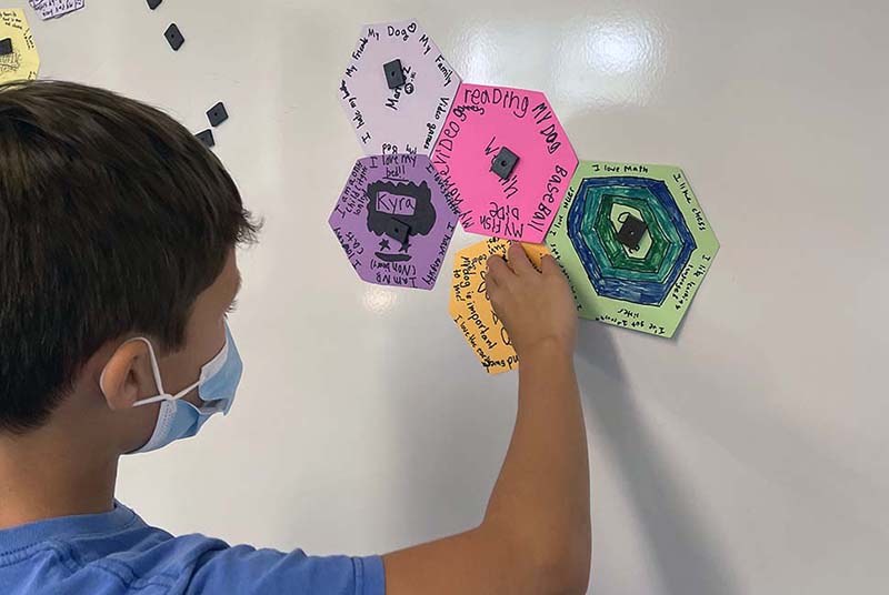 A Lower School student placing a bright orange hexagonal tile on a whiteboard next to some other brightly colored hexagonal tiles. Each of the tiles has handwritten notes on them.