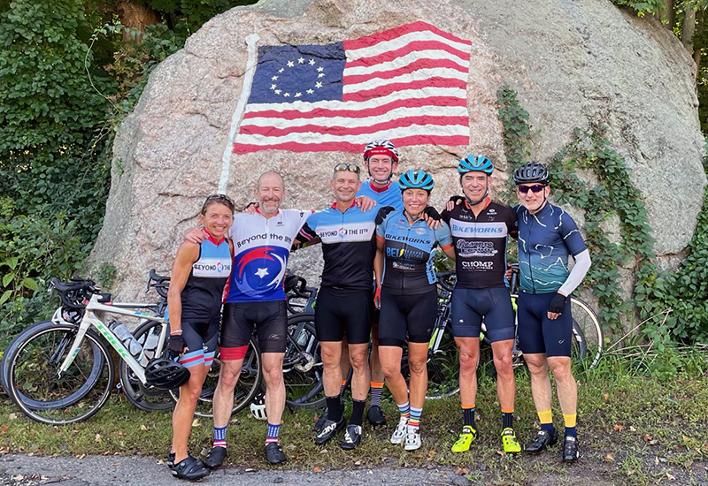 A group photo of Beyond the 11th bicycle riders wearing bike outfits and helmets. They are standing with their arms around one another and smiling at the camera. They are posing in front of a rock painted with the American flag featuring the circle of 13 stars representing the colonies.