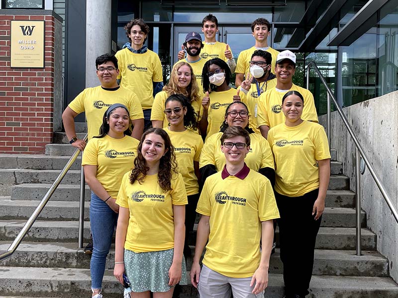 Breakthrough Providence staff pose on steps outside of a Wheeler School building and smile at the camera. They are all wearing yellow t-shirts that have a Breakthrough Providence logo on them.