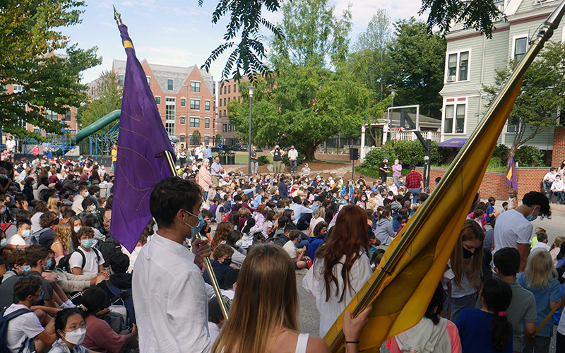 A photo of flag bearers walking into Convocation. One flag is purple and the other gold, and the flag bearers backs are to the camera. There is a large crowed seated outside in the background of the photo.