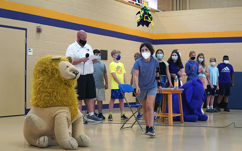 A photo of the Middle School sorting ceremony where a student walks towards the camera with the sorting hat handing overhead. Students are gathered in the background and a stuffed animal lion is in the foreground.