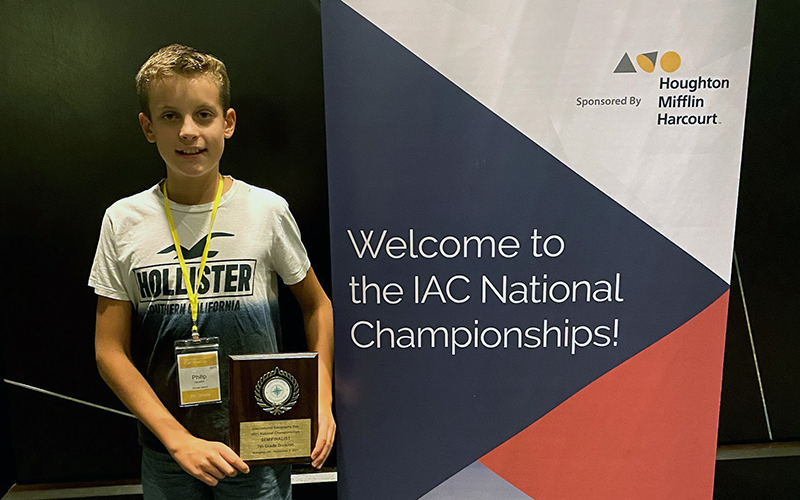 A photo of Philip Spradlin holding a semifinalist plaque and standing next to a banner than reads: "Welcome to the IAC National Championships!"