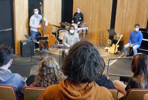 Students in the foreground talk with visiting jazz musicians on the stage in the background.}