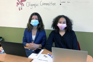 Model UN delegates sit at a table with their laptop computers. Olympic Committee is written on a whiteboard directly behind them.