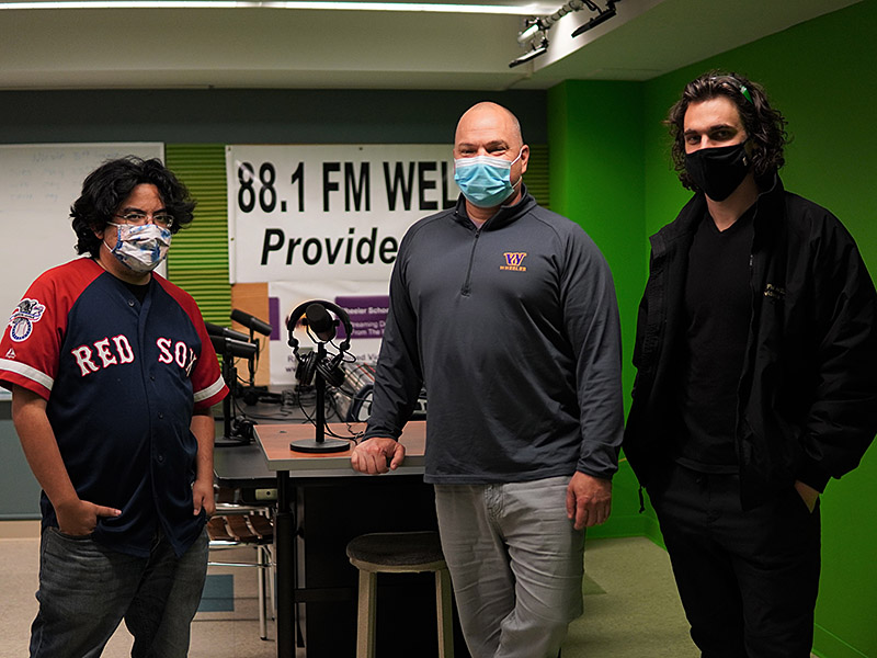 The WELH staff stands together in the WELH studio. They are wearing masks and looking at the camera.
