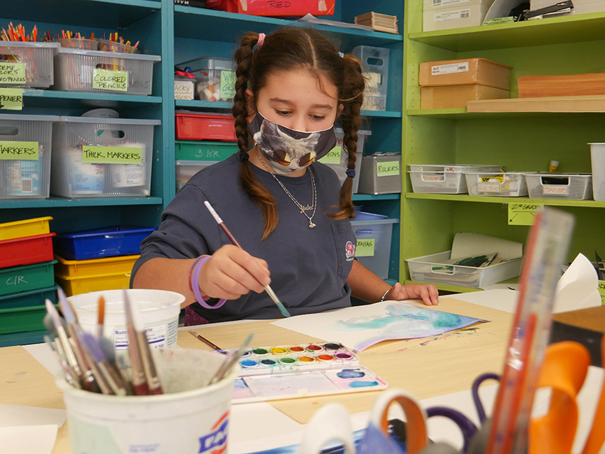 A 4th-grader student painters their holiday card. Art supplies are in the foreground of the picture.