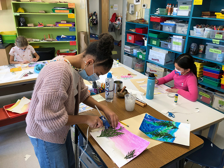 Students work on their holiday cards. One student in the foreground is using a hot glue gun to adhere pieces of nature to their piece while two students on either side in the background are painting.