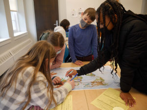 Students looking at a map together. There are white beans scattered across the map as part of a population exercise. Two of the students are sitting down at the table while two other students are standing.