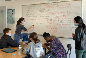 A group of students gathered by a whiteboard with notes written on it. One student is erasing something on the board while three others are seated a table and a fourth student stands nearby.