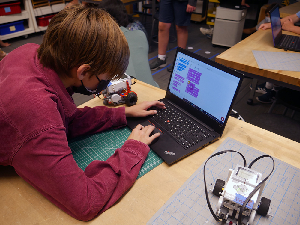 A Wheeler student working on laptop that is displaying programming commands for a robot while a small robot rests on the table next to them.