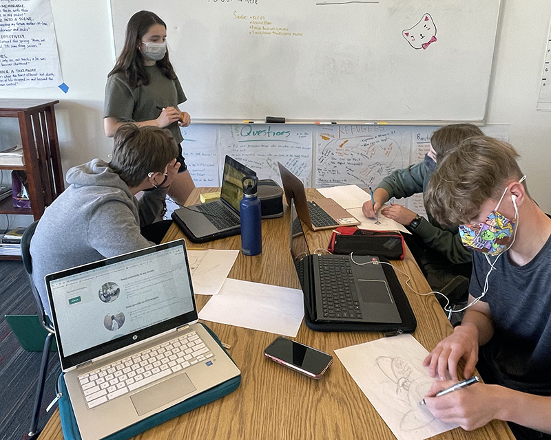 Students meeting to discuss their Cityside project. One student is standing by a whiteboard while three other students are seated at a table with laptop computers and drawings.