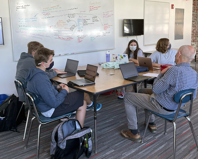 8th-graders meet with faculty member Joe Baer in the Cityside classroom. The students and faculty member are seated at a table--the faculty member's back is the camera) and there is a whiteboard with notes on the wall behind them.