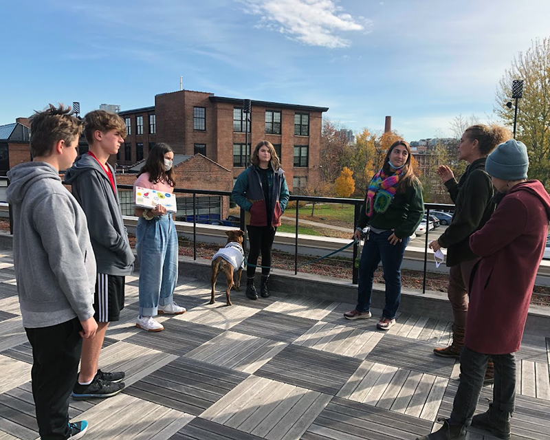 8th graders meet with their community partners outside on the deck of a building. They are standing in a circle while talking, and there is another building in the background behind them.