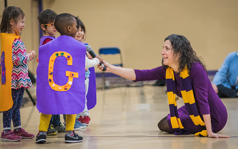 Head of School Allison Gaines Pell wearing a purple dress and yellow and purple scarf holds a microphone to Lower School students who are wearing purple and gold capes with letters on them.