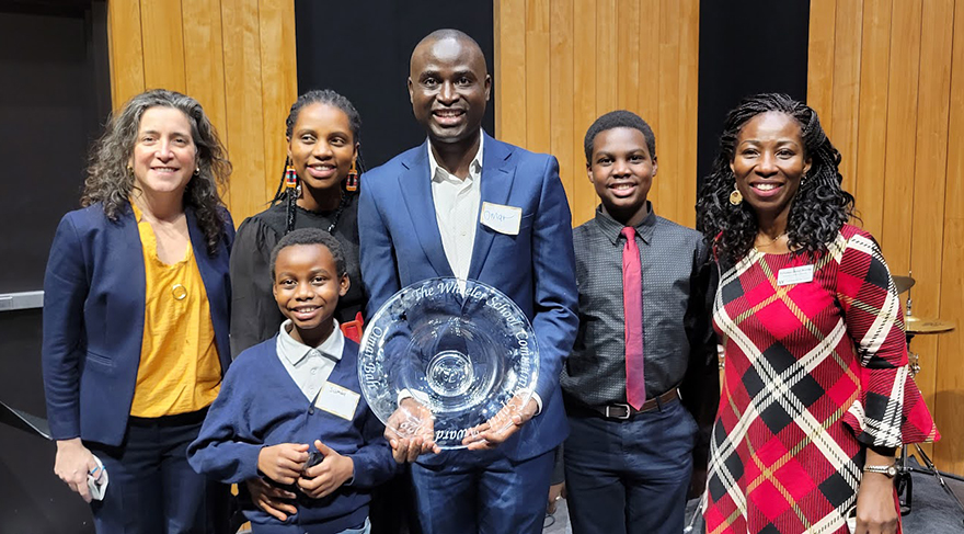 Omar Bah (center) holds the 2022 Wheeler Community Spirit Award and stands with family members, Head of School Allison Gaines Pell (far left), and Director of Unity and Diversity Princess Bomba (far right), inside Isenberg Auditorium.