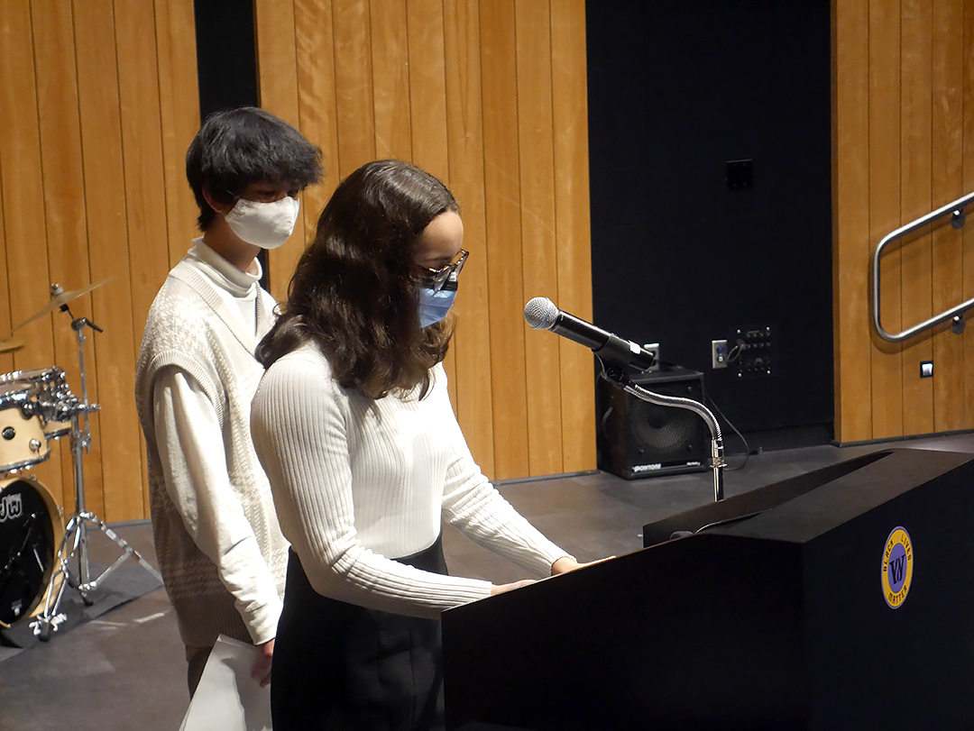 Two 8th-grade students stand a podium in Isenberg Auditorium with one speaking into a microphone while the other waits in the background.