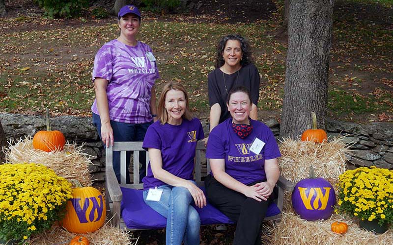 Allison Gaines Pell and three volunteers stand outside and smile at the camera and this year's Fall Fest. There are hay bales and pumpkins painted with the Wheeler "W" logo on either side of them.