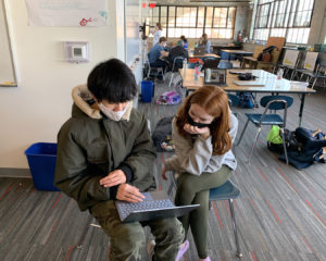 Charlotte Rampino ’26 and Philip Dang ’26 work together on their Cityside project in the WaterFire classroom. They are seated in chairs and looking at a laptop computer that Philip is holding. Other students are working in the background.
