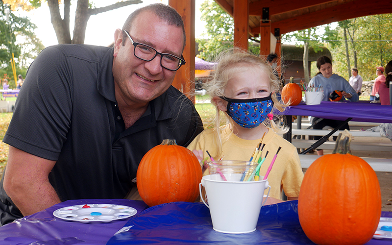 A parent and child painting pumpkins together while seated at a table outside.