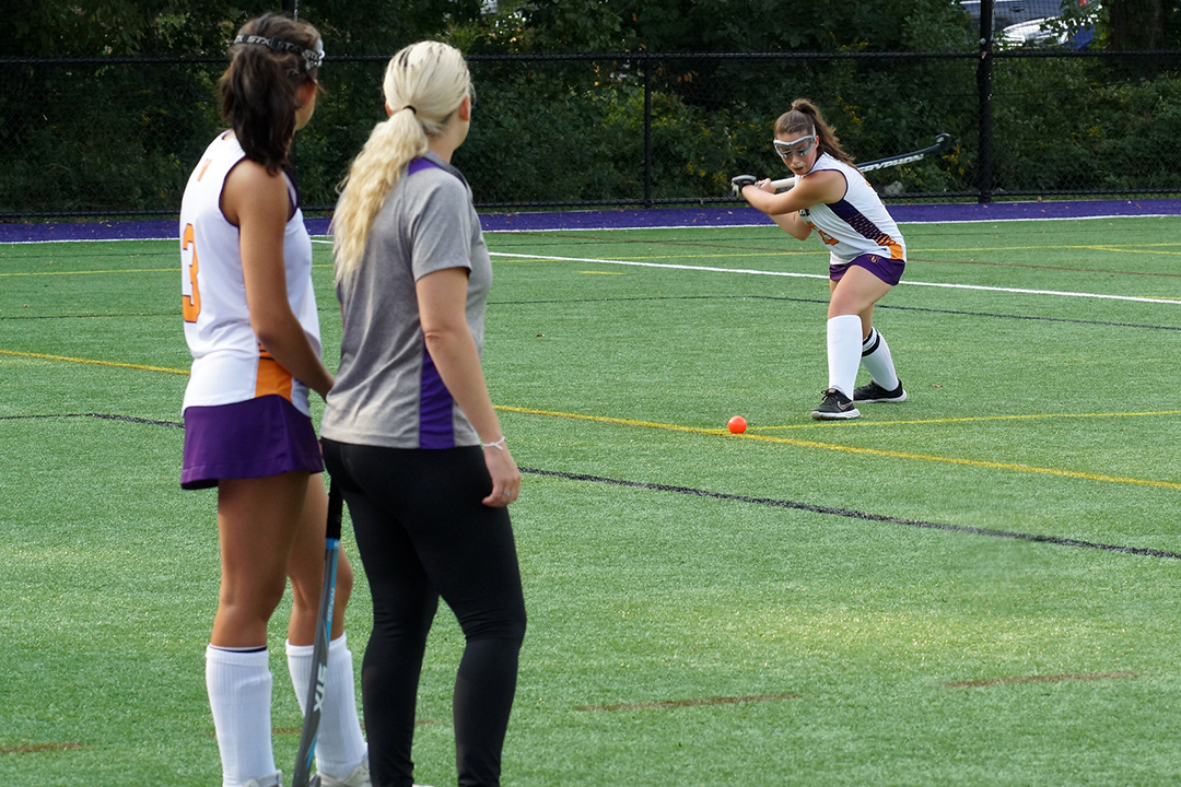 A field hockey coach and player watch another player swing at a ball.
