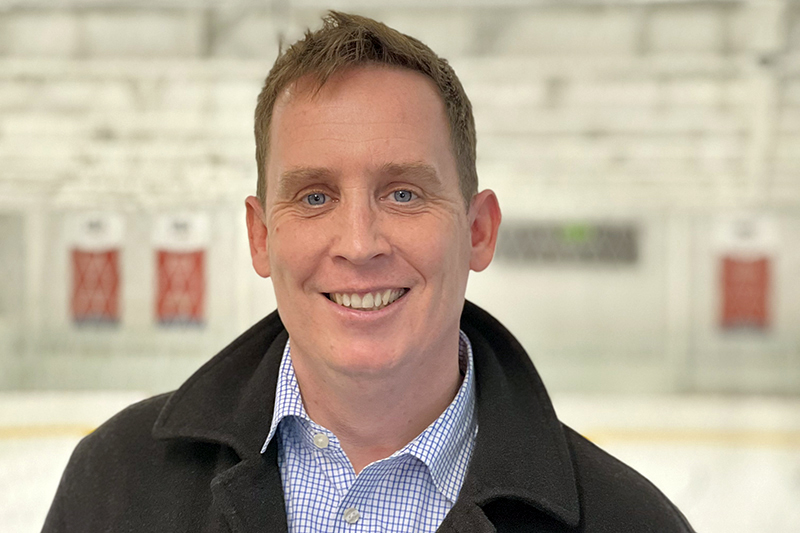 Photo of Ian Moorhouse smiling at the camera. Though the background is blurry, he appears to be standing in an ice rink and appears from the chest up.