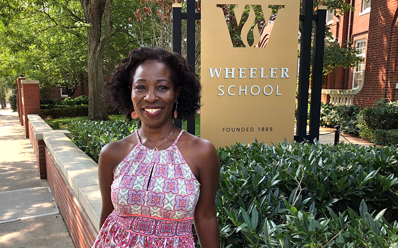 Princess Bomba stands outside and smiles at the camera. She is standing in front of the Wheeler School sign at the front entrance to campus.