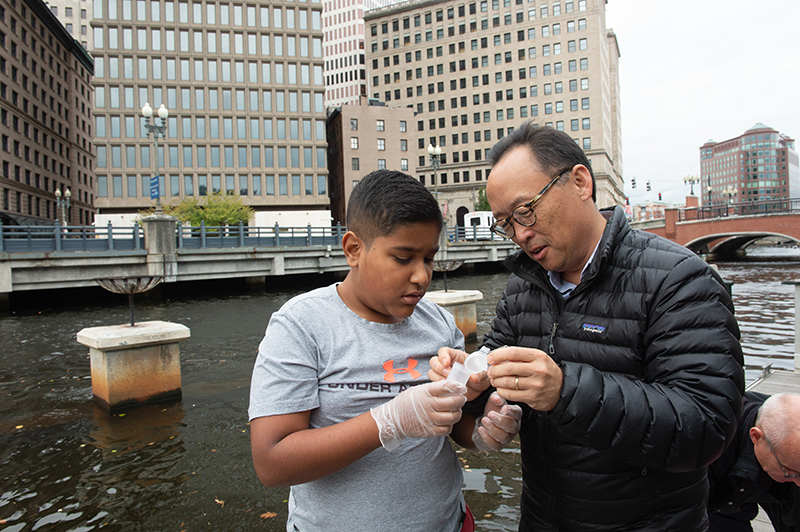 Young Un and a student stand by the water in downtown Providence and look at a sample of something in a test tube.