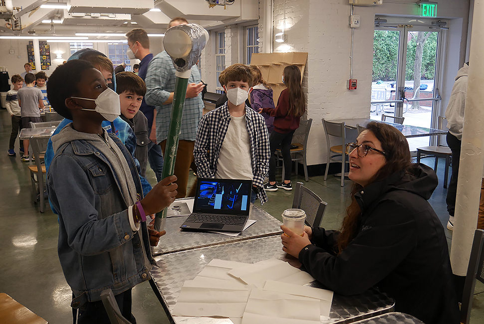 A student raises a tool they created alongside fellow students as a teacher watches the demonstration while seated.