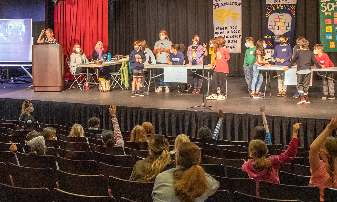 Students in the audience raise their hands at the 2022 Battle of the Books while other student teams are gathered together on stage.