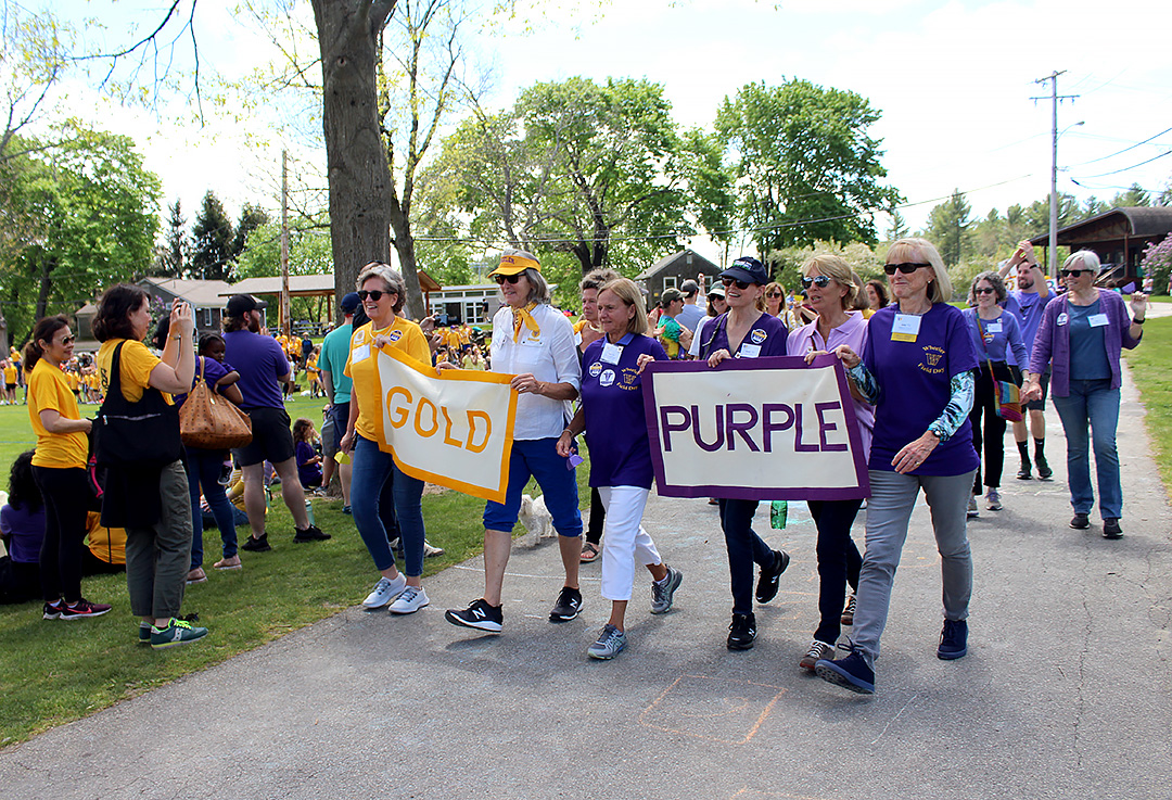 A group of alumni carry signs while wearing purple and gold shirts in front a crowd at Wheeler Farm.
