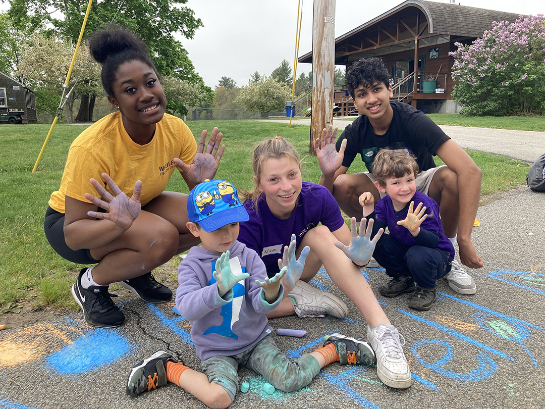 A trio of Upper School students hold up hands covered in colored chalk while sitting with two younger Nest students on top of chalk drawings on a road at Wheeler Farm.