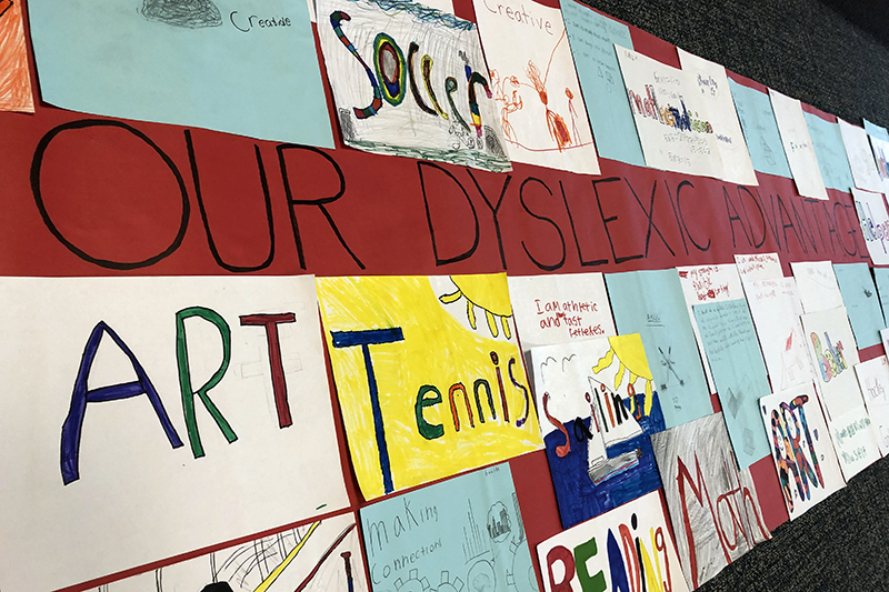 A red banner with a handwritten title in the middle that reads: "Our Dyslexic Advantage." The title is surrounding by individual rectangles of paper that say different words that signify different strengths, such as "art," "tennis," and "math."