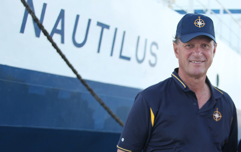 Dr. Ballard stands in front of blue and white ship with "Nautilus" written on the side in blue uppercase letters.