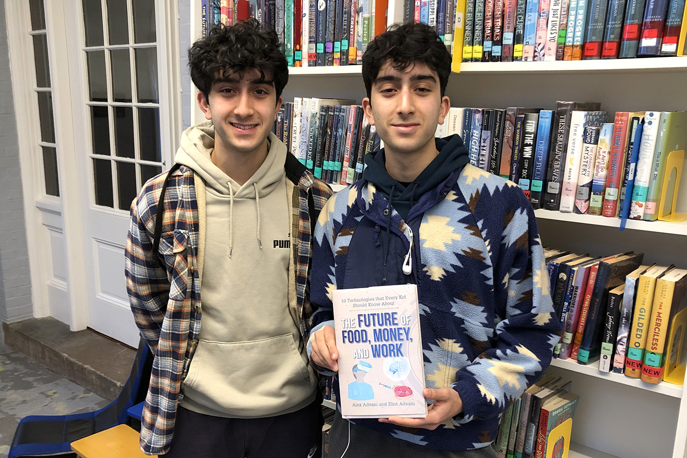 A photo of Alex and Eliot Advani posing while holding their book. Books line a series of shelves behind them.