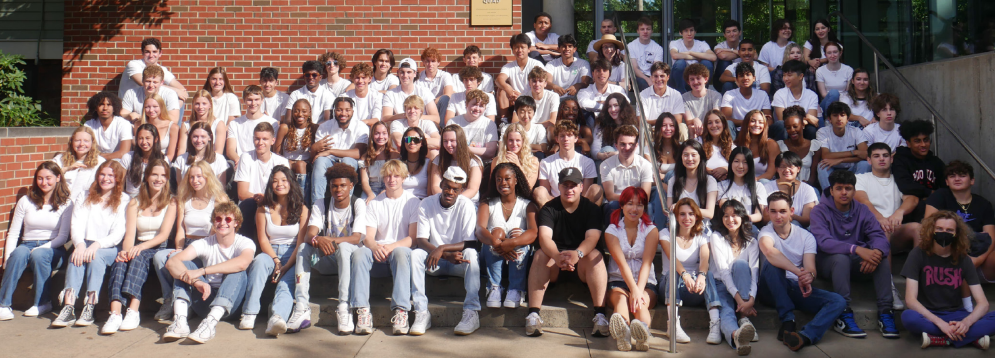 A group photo of the Class of 2023 sitting together on steps in Miller Quad. Most of the students are wearing white t-shirts.