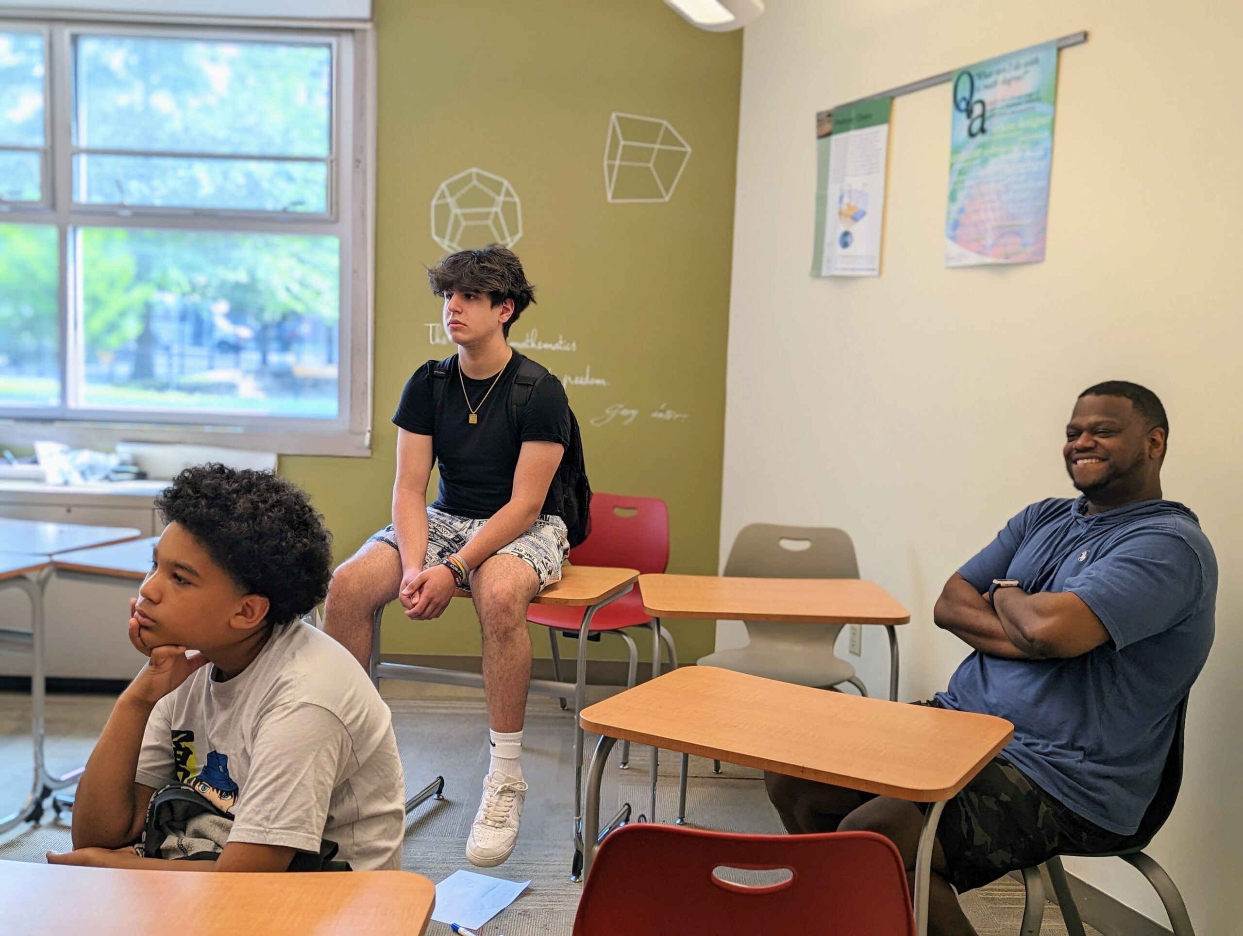 Photo of Mr. Davis, including two students, sitting down and listening to students who are presenting. Mr. Davis has a smile on his face.